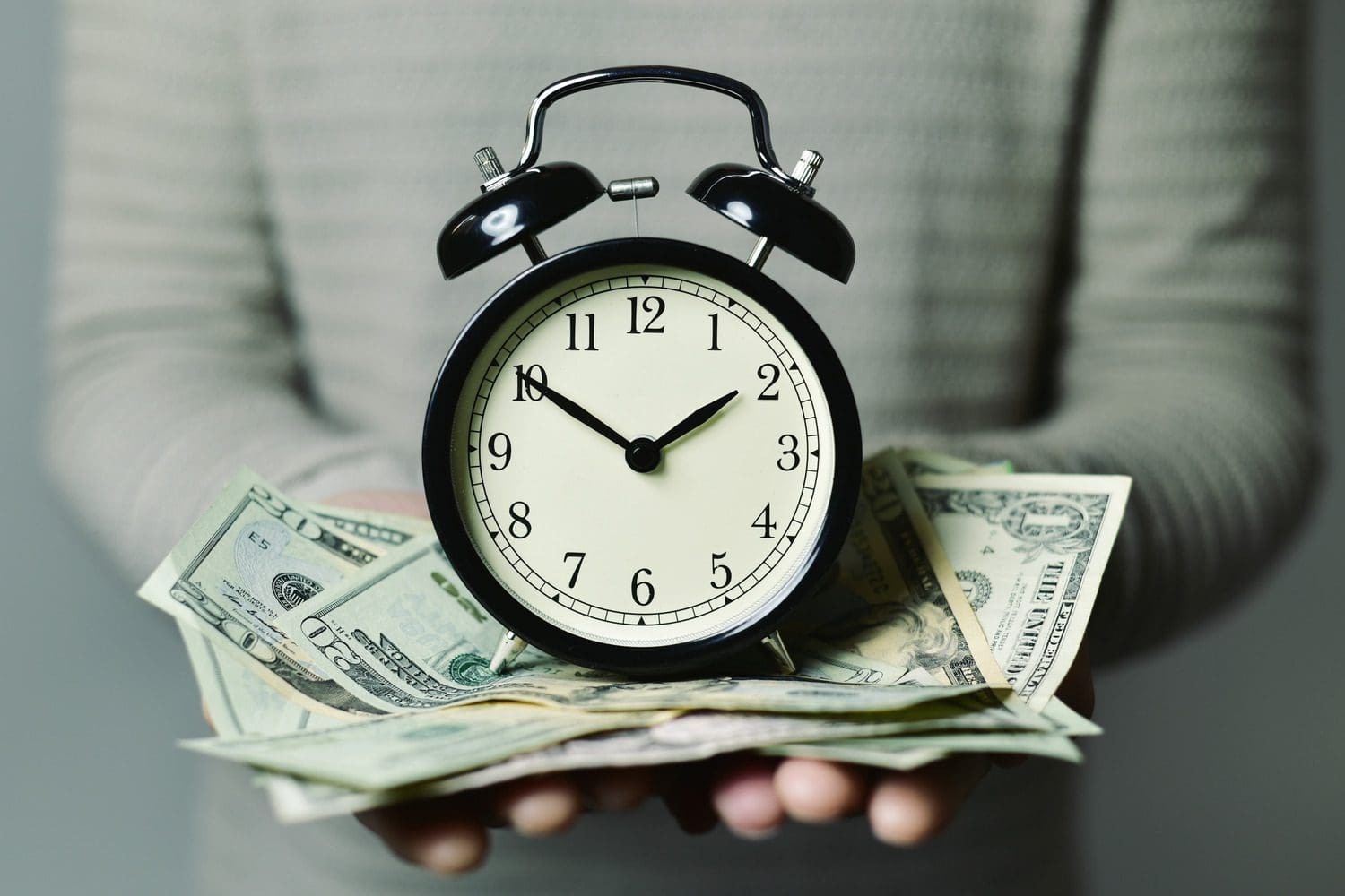 An analog alarm clock sitting on a stack of U.S. dollar bills representing the concept of financial vesting and time-based incentives.