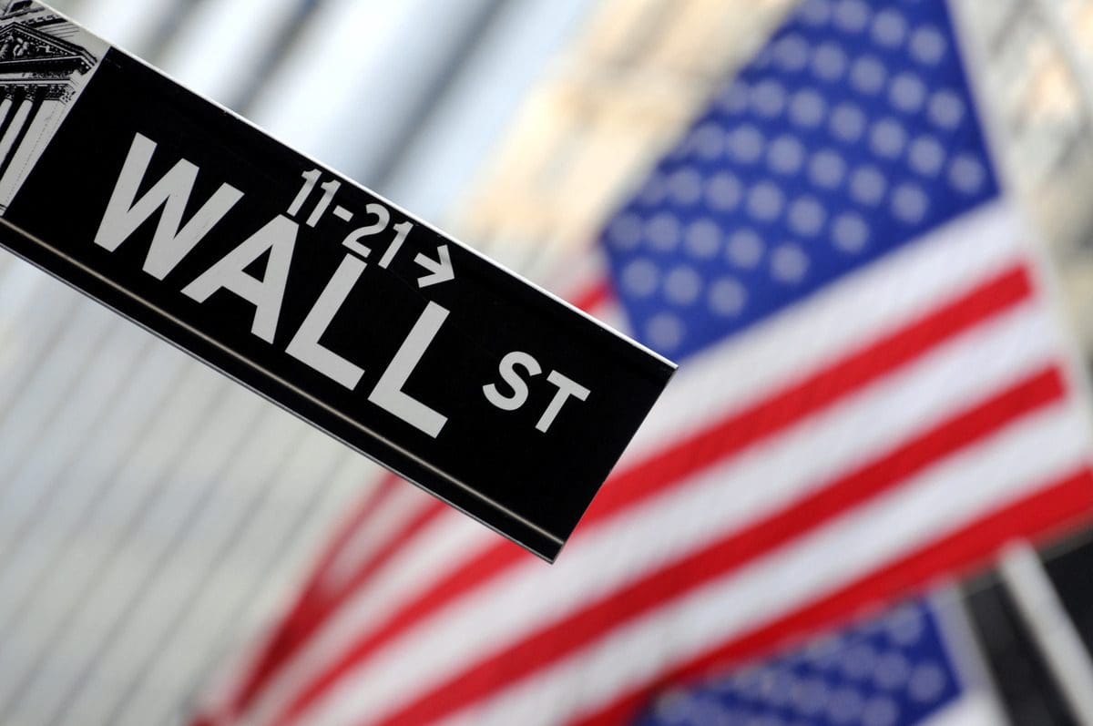A street sign for Wall Street with a blurred American flag in the background, representing the U.S. financial system.