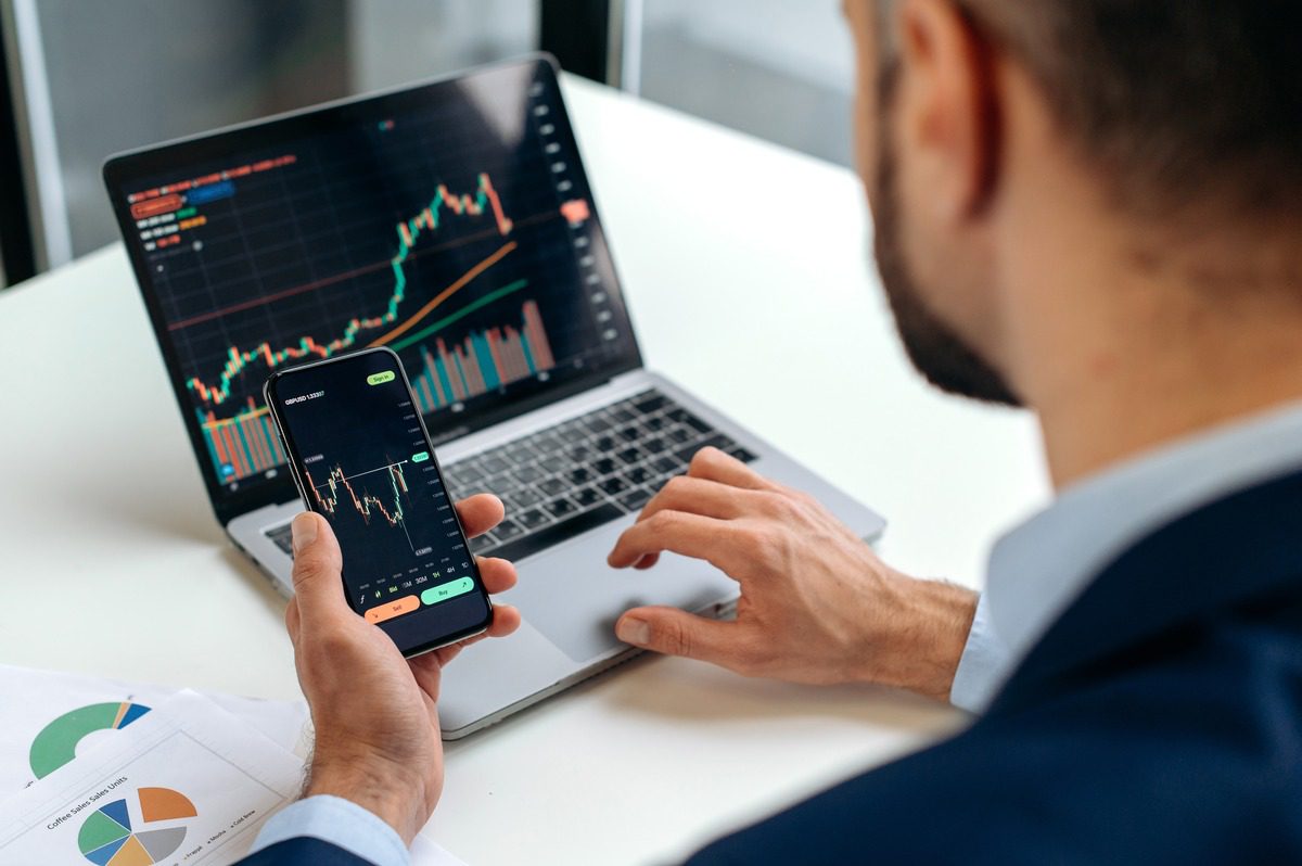 Person monitoring cryptocurrency market trends and candlestick charts on a laptop and smartphone.